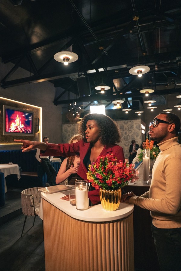 keepers-of-the-door-01 In a warmly lit restaurant, a woman in a red blazer points, standing next to a man in a beige ribbed sweater.