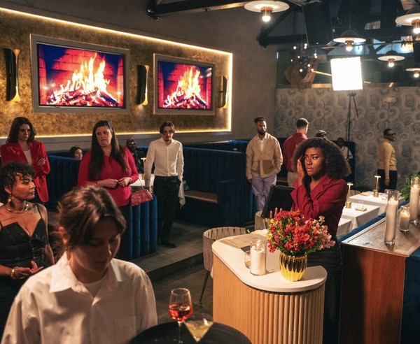 keepers-of-the-door-03 A hostess in a red jacket raises her hand to her face amid a bustling restaurant dining room decorated with glowing fireplace screens.