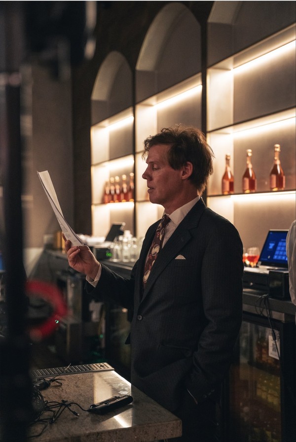 keepers-of-the-door-04 A man in a dark suit reads from a sheet of paper in front of the restaurant bar, surrounded by illuminated bottles.