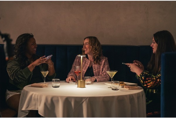 keepers-of-the-door-05 Three smiling friends seated at a table with cocktails, in a dimly lit restaurant setting.